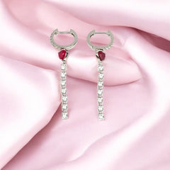 A pair of silver hoop earrings with heart-shaped dangles and zircon stones, displayed on a pink satin background.
