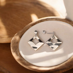 A pair of silver rhombus dangler earrings placed on a wooden surface with a shadow cast over them.