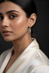 A close-up of a person's ear wearing a silver huggie hoop earring with a dangling heart-shaped charm and clear crystal accents.