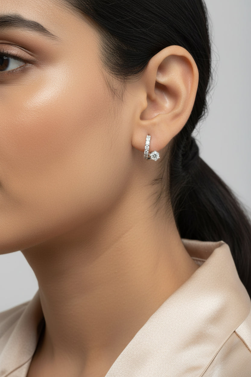 A close-up of a person's ear wearing a small silver huggie hoop earring adorned with clear stones.