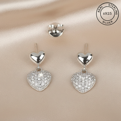 Silver heart-shaped earrings with sparkling details on a beige fabric background, featuring 'Real Sterling Silver' certification.