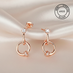 Rose gold earrings with clear gemstones on a beige background, featuring 'Real Sterling Silver 925' stamp.