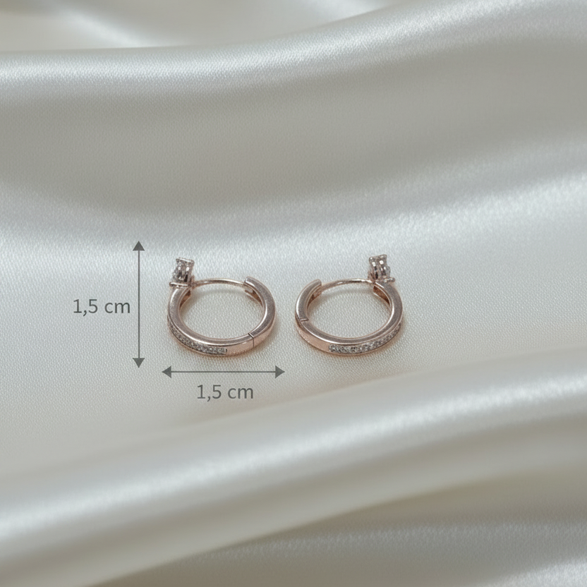 A pair of small rose gold hoop earrings with a single clear gemstone on each earring, displayed on draped white satin fabric.