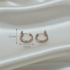 A pair of small rose gold hoop earrings with a single clear gemstone on each earring, displayed on draped white satin fabric.