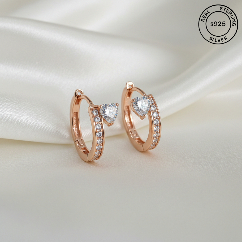 A pair of rose gold huggie hoop earrings with heart-shaped clear gemstones and pavé accents, displayed on a draped white satin fabric. A circular 'REAL STERLING S925 SILVER' logo is visible in the top right corner.