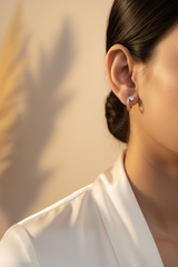 A close-up of a person's ear wearing a small rose gold hoop earring with a clear heart-shaped crystal and a pavé-set crystal bar.
